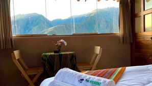 Habitación de hotel con una cama, dos sillas de madera, una mesita con un jarrón de flores y vistas a montañas a través de la ventana.