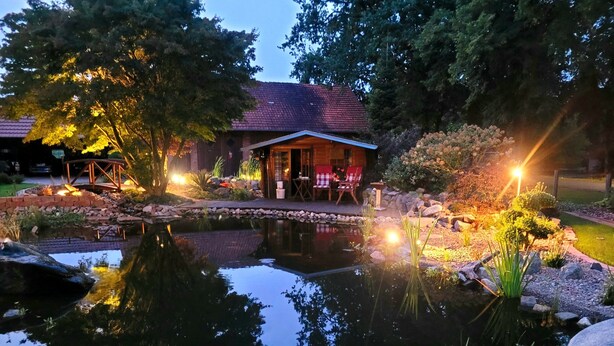A pond with a wooden bridge, a small building with a red roof, and outdoor seating by the water.