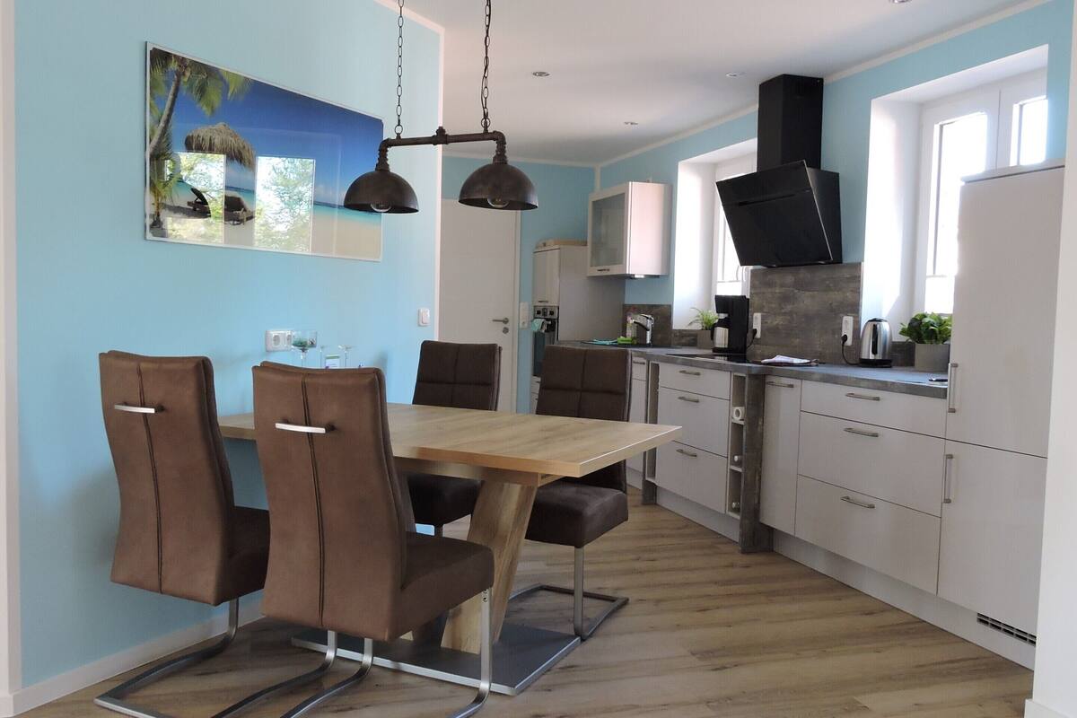 A modern kitchen with a wooden dining table and chairs, a built-in oven, and a large window.