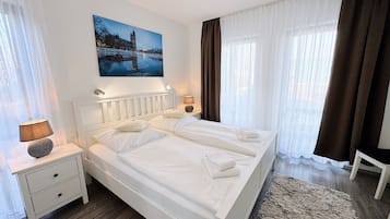 A hotel room with a white bed, two bedside tables, a nightstand, a lamp, a painting of a cityscape, and a view of a building through the window.