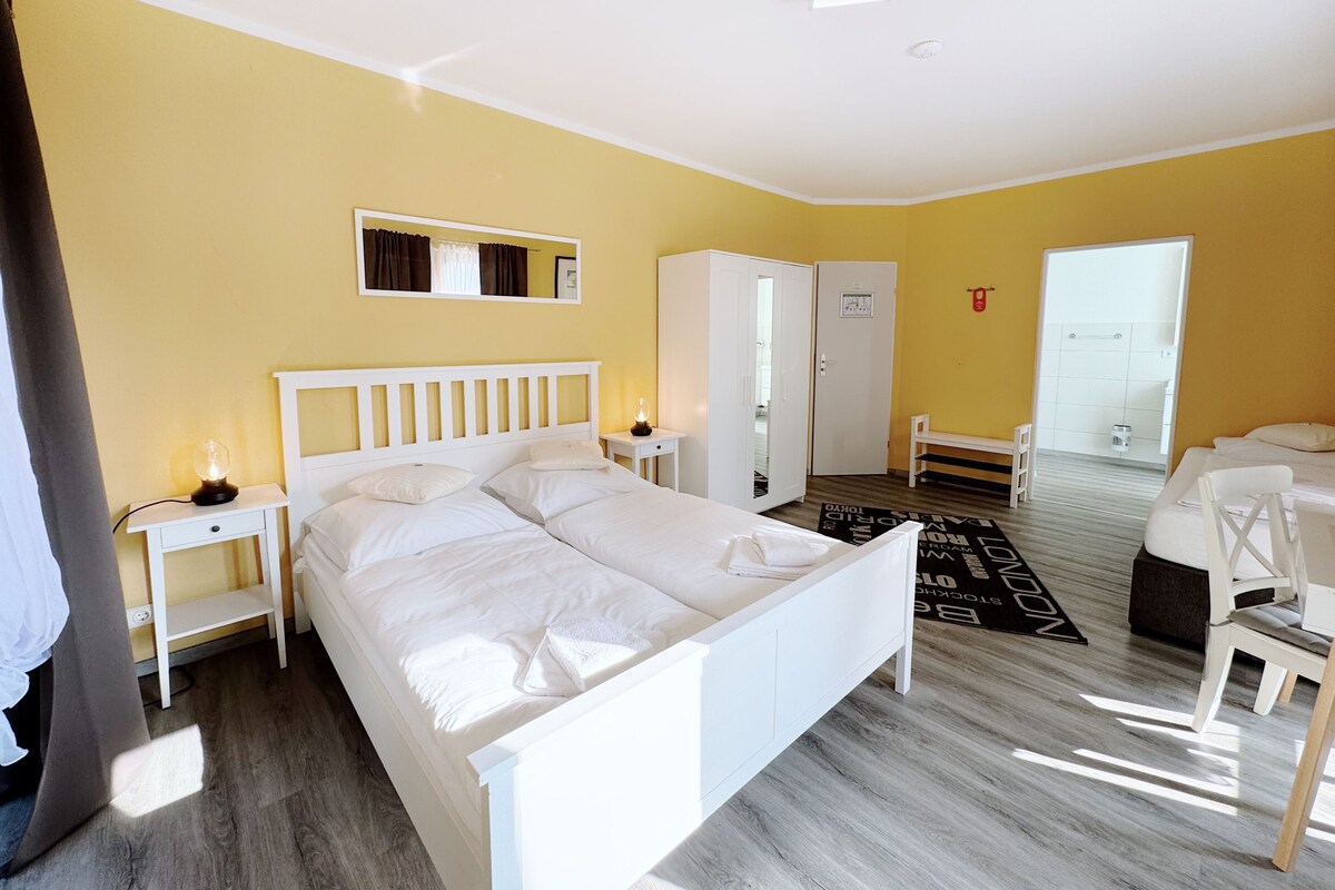 A hotel room with a double bed, a bedside table with a lamp, a wardrobe, a bench, and a door leading to a bathroom.