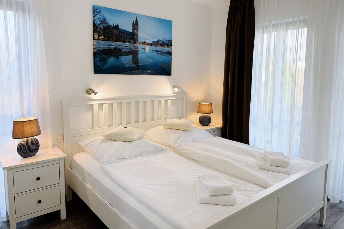 A neatly made bed with white linens and pillows, a bedside table with a lamp, and a painting of a cityscape above the bed.