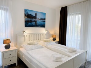 A neatly made bed with white linens and pillows, a bedside table with a lamp, and a painting of a cityscape above the bed. - Regiohotel Wittekind Burg (Burg)