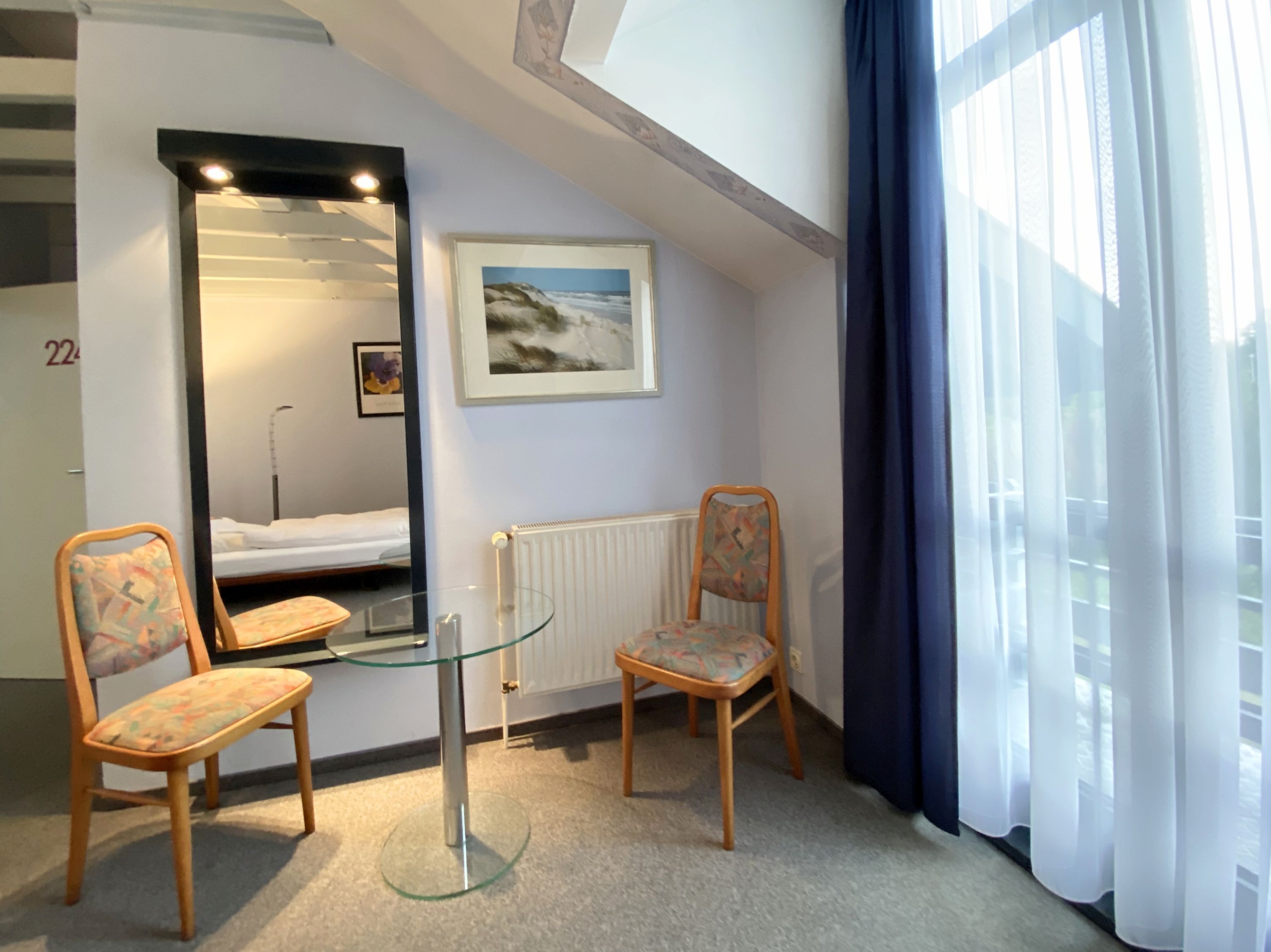 Quarto de hotel com cama, duas cadeiras, mesa de vidro, radiador, quadro emoldurado e janela com cortinas.