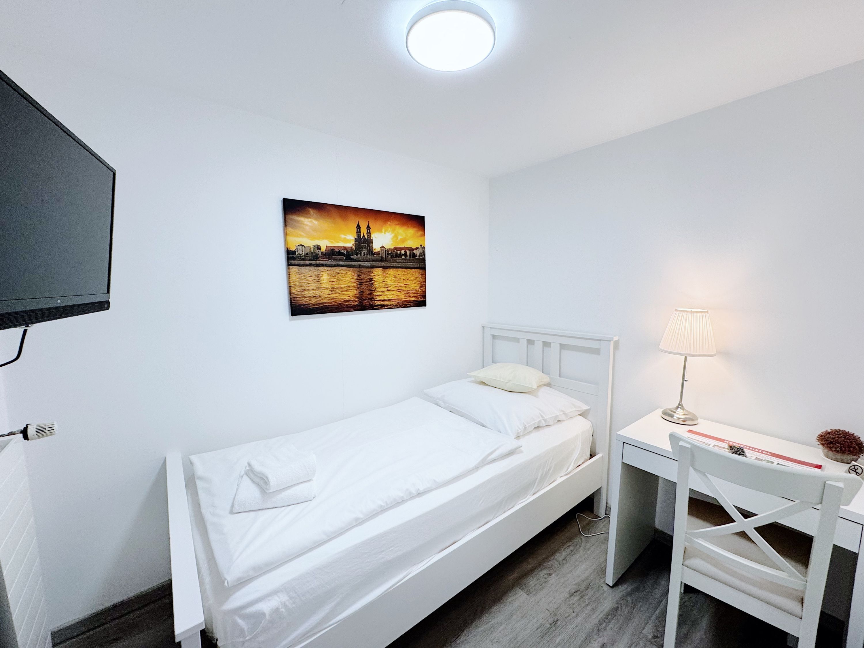 A single bed with white bedding, a desk with a lamp, and a painting of a sunset over a cityscape on the wall.