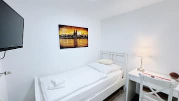 Ein Einzelbett mit weißer Bettwäsche, ein Schreibtisch mit Lampe und ein Gemälde, das einen Sonnenuntergang über einer Stadt zeigt.