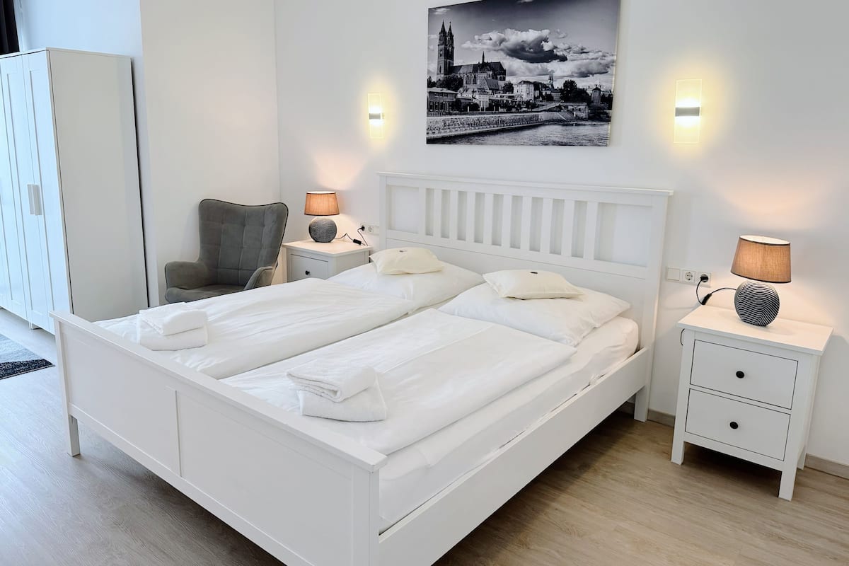 A modern bedroom with a white bed, two bedside tables with lamps, a chair, and a large cityscape painting on the wall.