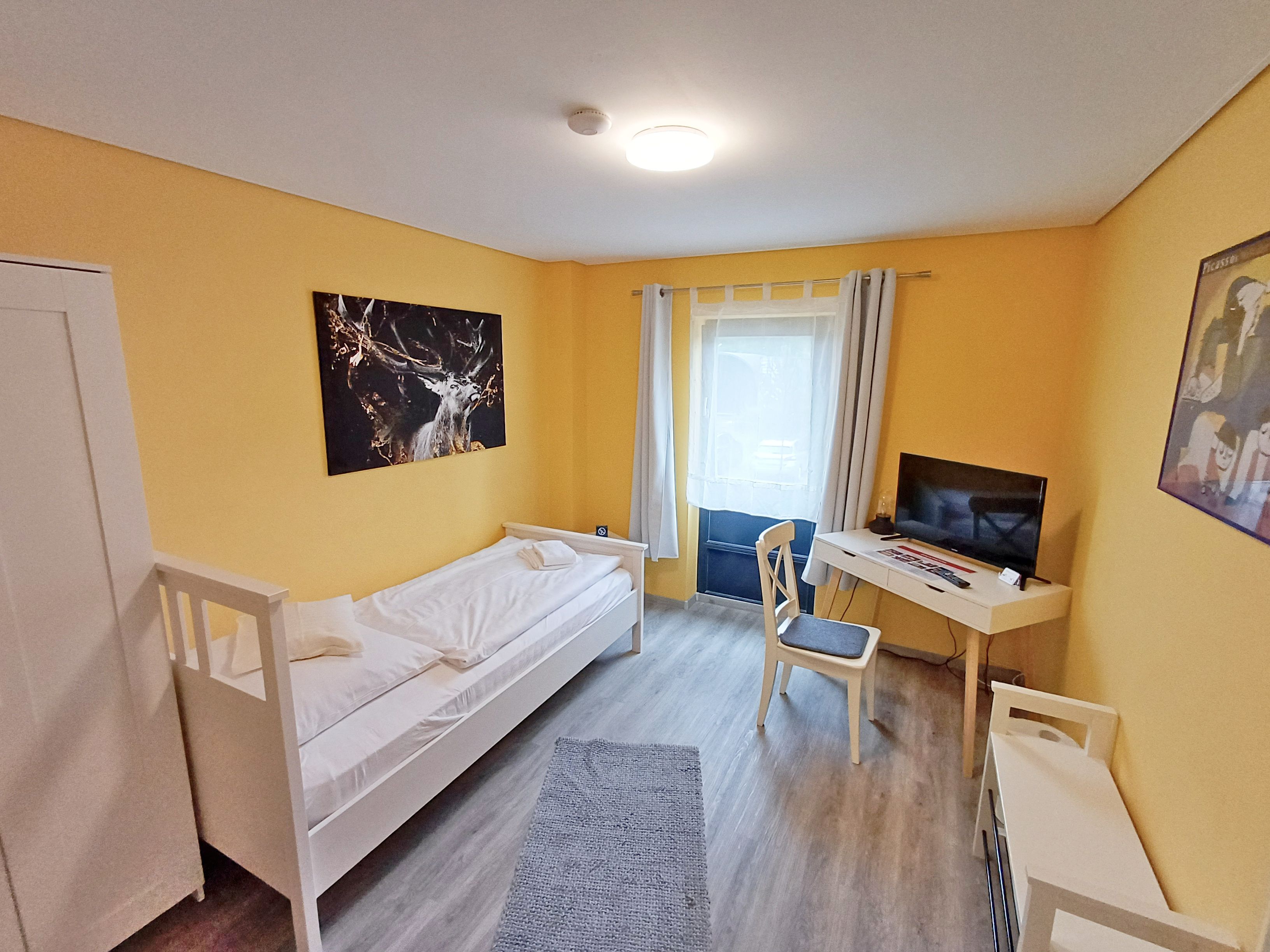 Ein kleiner, heller Raum mit einem Bett, einem Schreibtisch mit Computer und einem fenster mit Vorhängen.