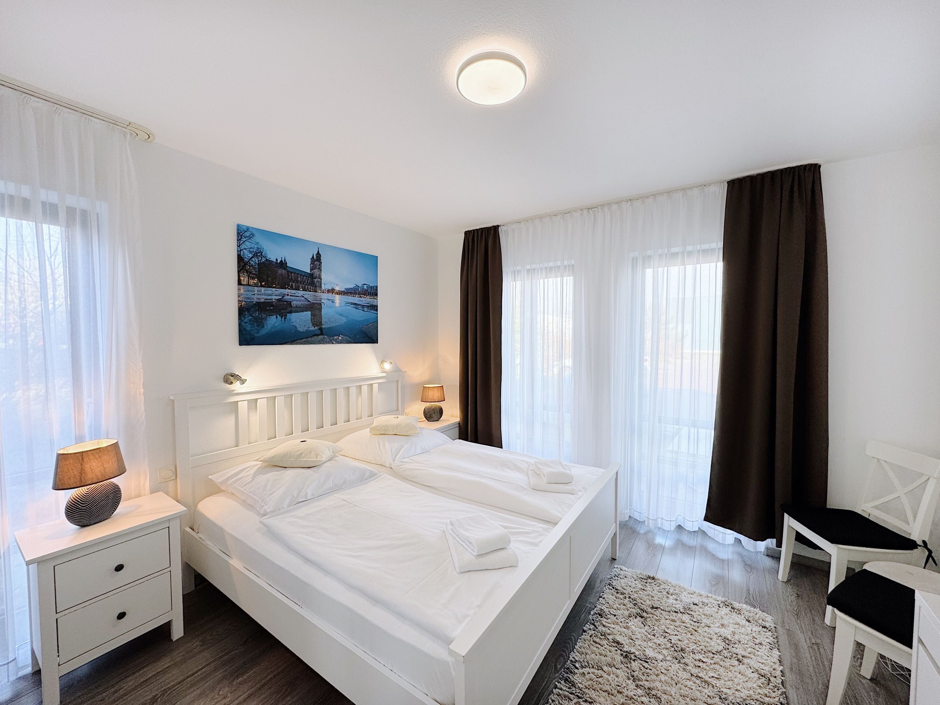 A hotel room with a double bed, two bedside tables with lamps, a chair, a large window with curtains, and a painting of a cityscape.