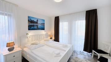A hotel room with a double bed, two bedside tables with lamps, a chair, a large window with curtains, and a painting of a cityscape.