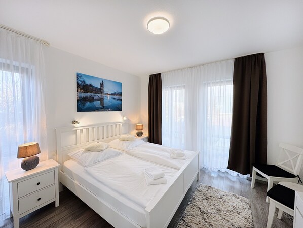 Et hotelværelse med en dobbeltseng, to sengeborde med lamper, en stol, et stort vindue med gardiner og et maleri af et bybillede.