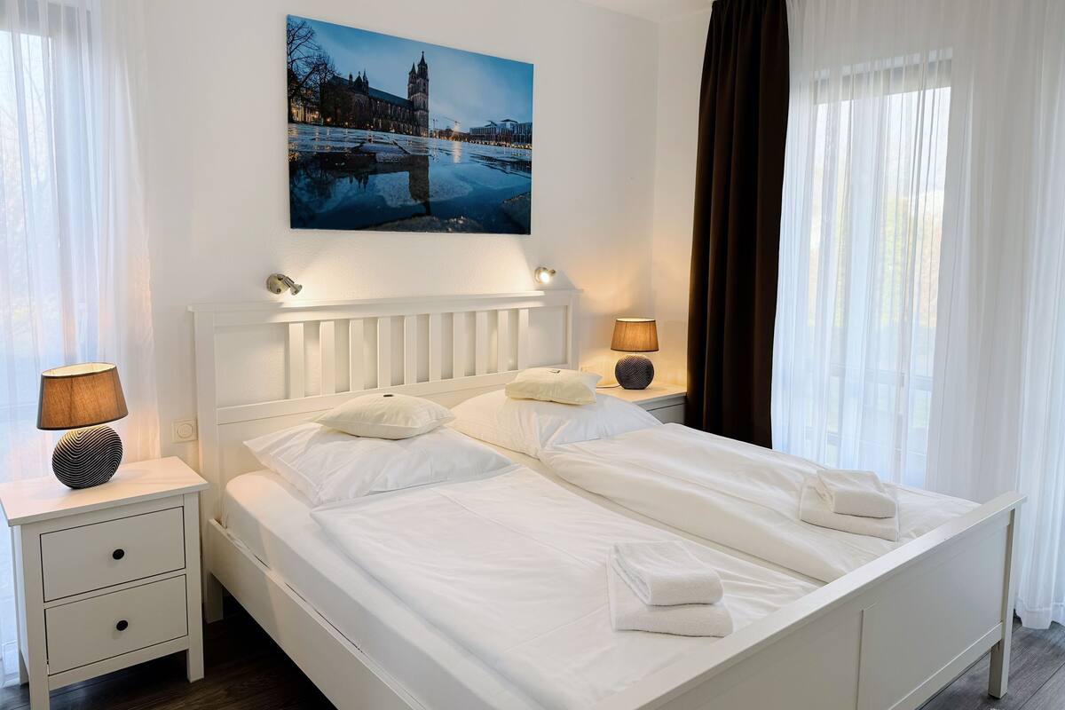 A neatly made bed with white linens and pillows, a bedside table with a lamp, and a painting of a cityscape above the bed.