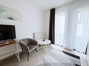 Ein modernes, minimalistisches Wohnzimmer mit Fernseher, einem grauen Sessel, einem kleinen weißen Tisch und einem großen Fenster mit transparenten Vorhängen.