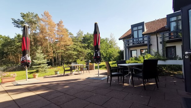 An outdoor patio area with tables and chairs, two umbrellas, and a building with a tiled roof in the background. - Regiohotel Wittekind Burg (Burg)