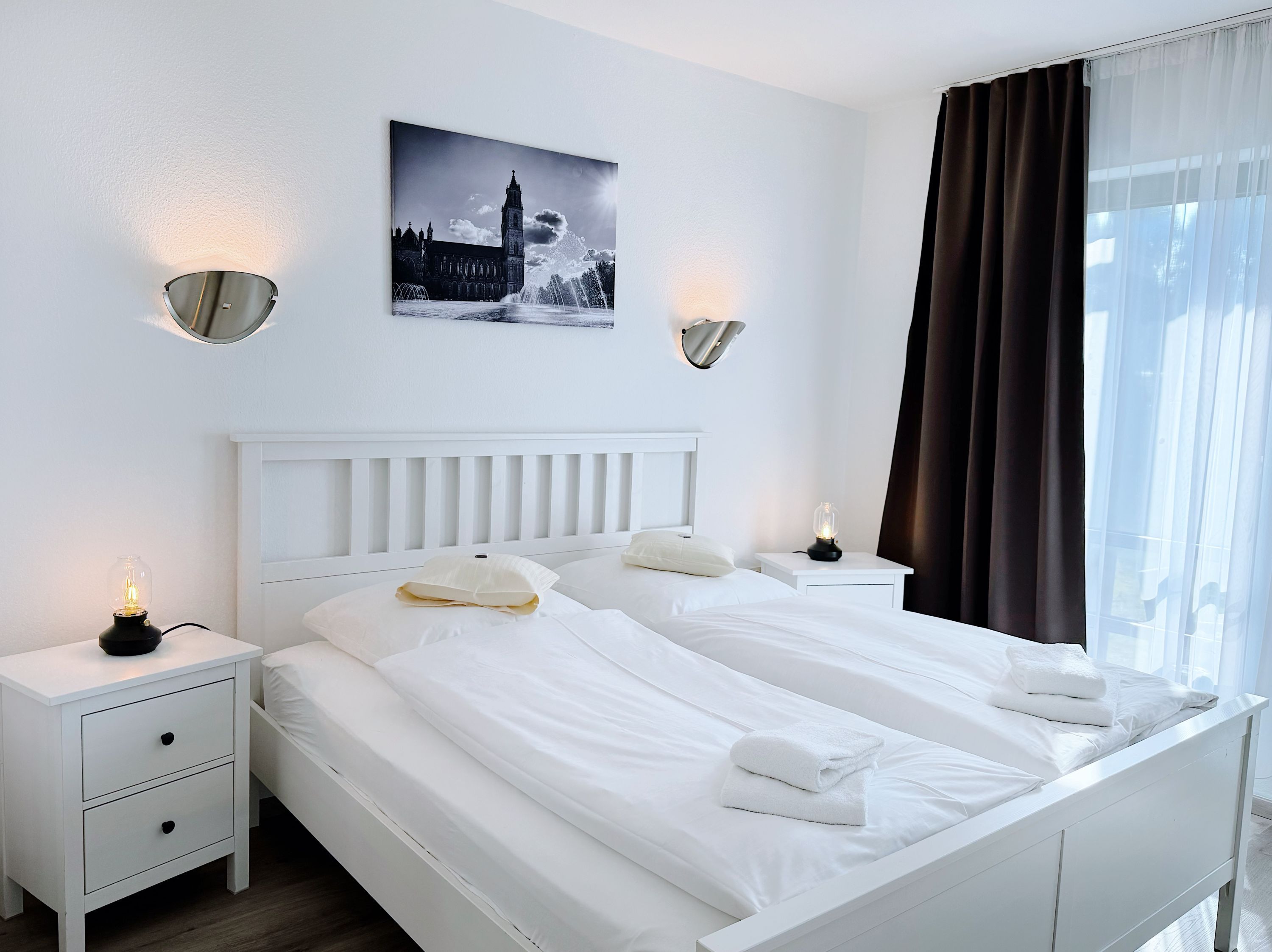 Ein Hotelzimmer mit zwei Betten, weißer Bettwäsche, einem Nachttisch mit Lampe, einem Wandbild und einem Fenster mit Vorhängen.