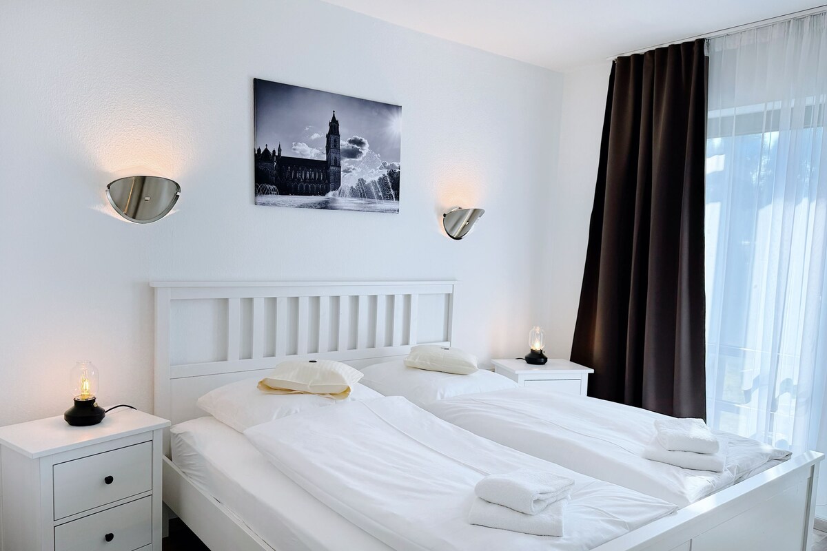 A hotel room with two beds, white bedding, a nightstand with a lamp, a wall-mounted picture, and a window with curtains.
