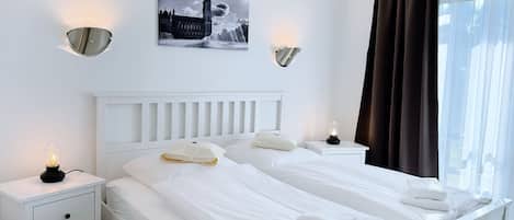 Habitación de hotel con dos camas, ropa de cama blanca, una mesita de noche con lámpara, un cuadro en la pared y una ventana con cortinas.