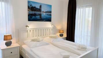 Una cama bien hecha con sábanas y almohadas blancas, una mesita de noche con una lámpara, y un cuadro de un paisaje urbano sobre la cama.