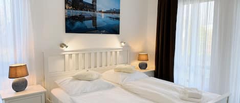 Een net opgemaakt bed met witte lakens en kussens, een nachtkastje met een lamp, en een stadsgezichtschilderij boven het bed.