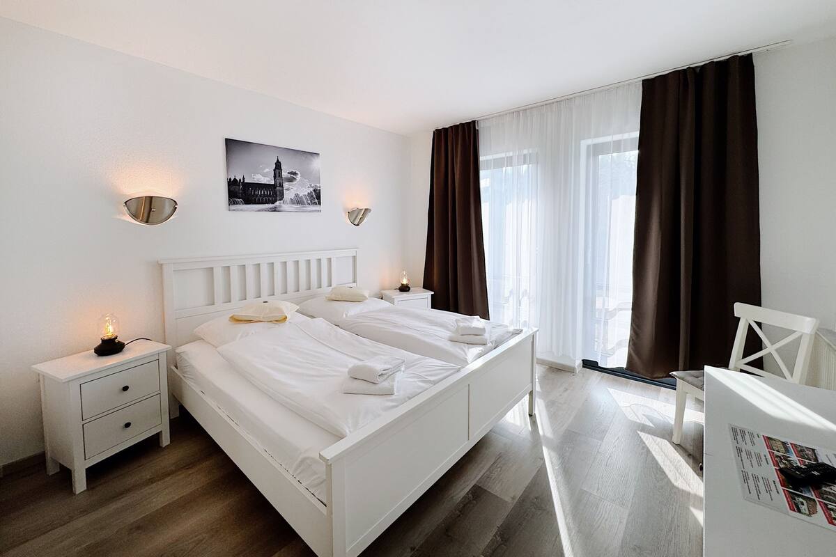 A hotel room with a white bed, two bedside tables, a desk, and a chair. There are two wall-mounted light fixtures and a framed picture on the wall.