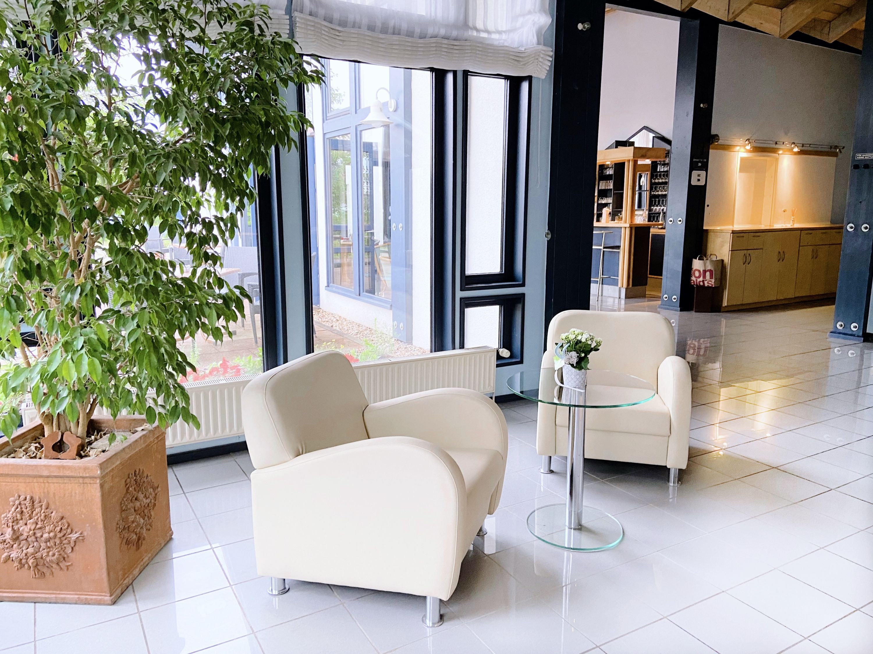 En modern hotellobby med ett stort träd i en kruka, två vita fåtöljer, ett glasbord och utsikt över utomhusområdet genom stora fönster.