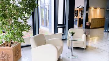 En modern hotellobby med ett stort träd i en kruka, två vita fåtöljer, ett glasbord och utsikt över utomhusområdet genom stora fönster.
