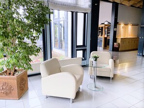 Un moderno vestíbulo de hotel con un gran árbol en maceta, dos sillones blancos, una mesa de vidrio y vistas al área exterior a través de amplias ventanas.