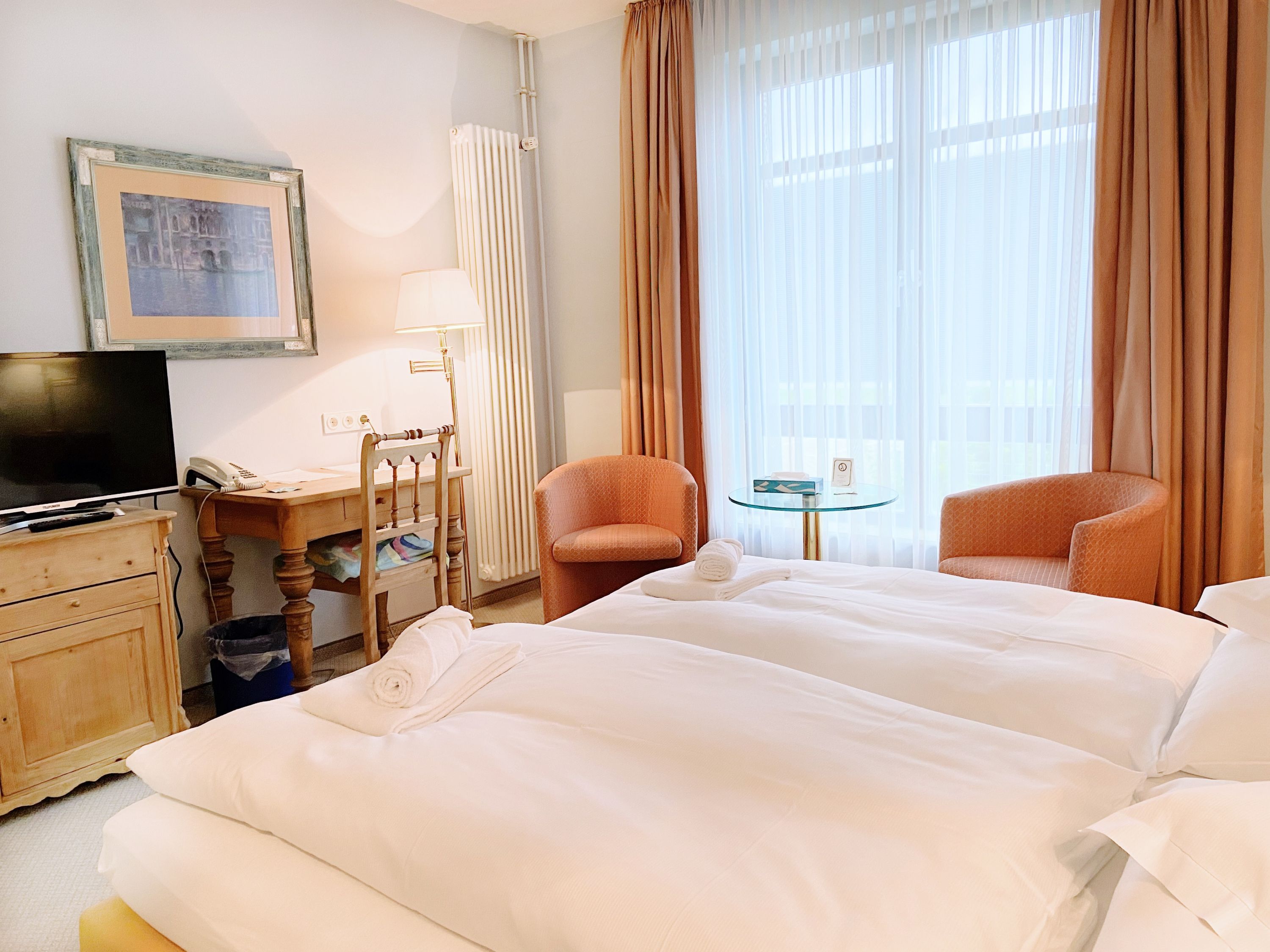 Habitación de hotel con dos camas, un escritorio con lámpara, una silla, una mesita y un televisor.