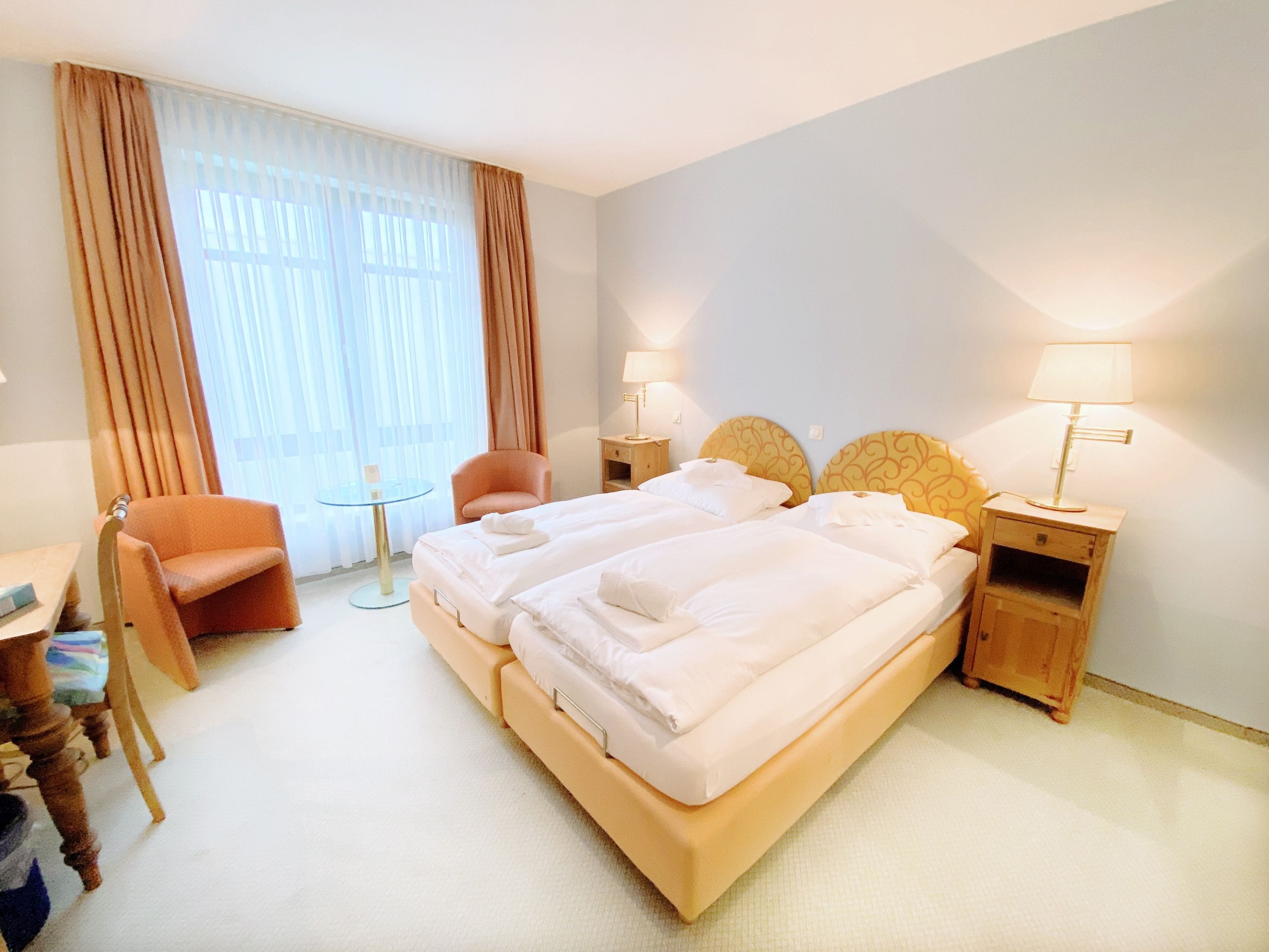 Quarto de hotel com uma cama de casal, duas mesas de cabeceira com luminárias, uma cadeira, uma mesinha redonda e uma escrivaninha com cadeira.