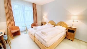 Quarto de hotel com uma cama de casal, duas mesas de cabeceira com luminárias, uma cadeira, uma mesinha redonda e uma escrivaninha com cadeira.