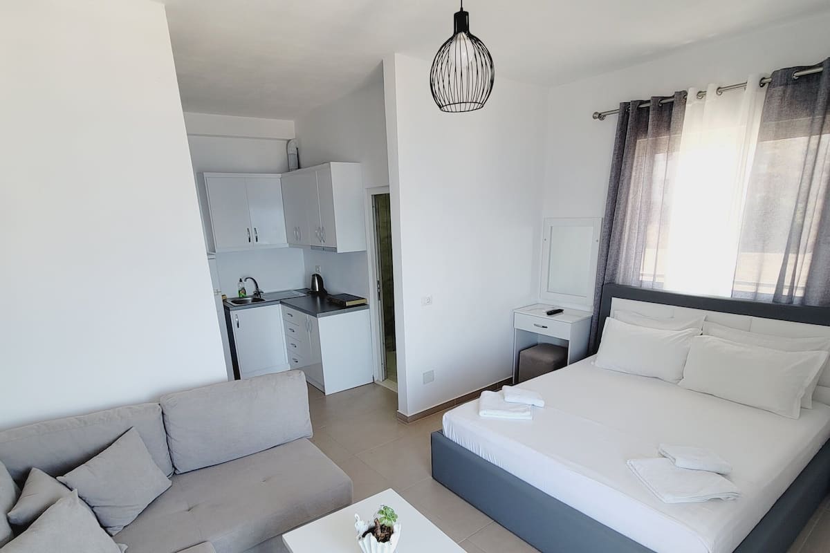 A modern, well-lit room with a grey sofa, a white coffee table, a large bed with white linens, and a kitchen area in the background.