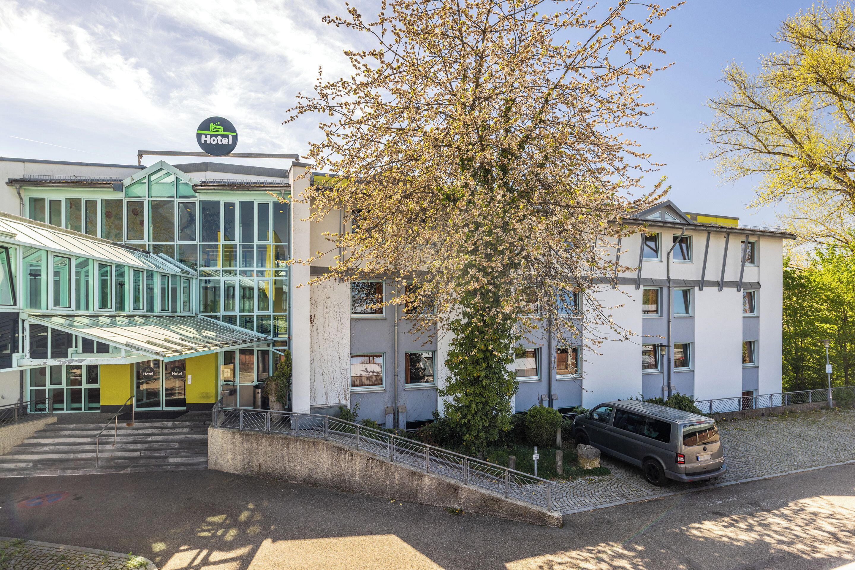 Et hotel med en moderne glasseftergang, parkeringsområde og et træ foran.