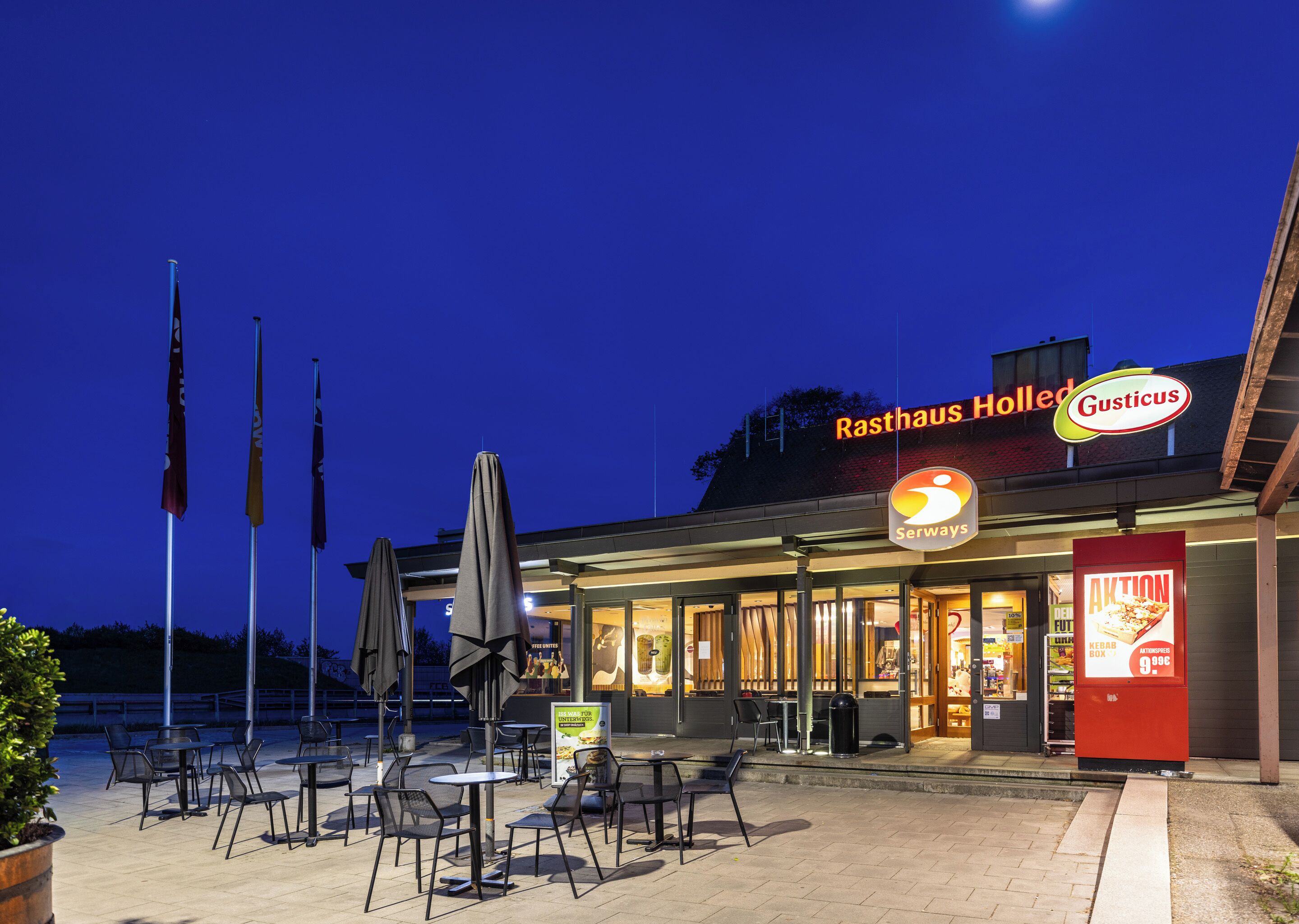 An outdoor dining area with tables and chairs, flags, and a restaurant named 'Rasthaus Holled' with a 'Gusticus' sign.