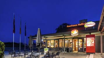 An outdoor dining area with tables and chairs, flags, and a restaurant named 'Rasthaus Holled' with a 'Gusticus' sign.