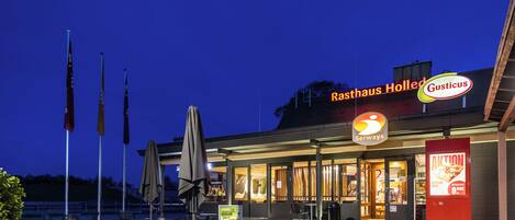 An outdoor dining area with tables and chairs, flags, and a restaurant named 'Rasthaus Holled' with a 'Gusticus' sign.
