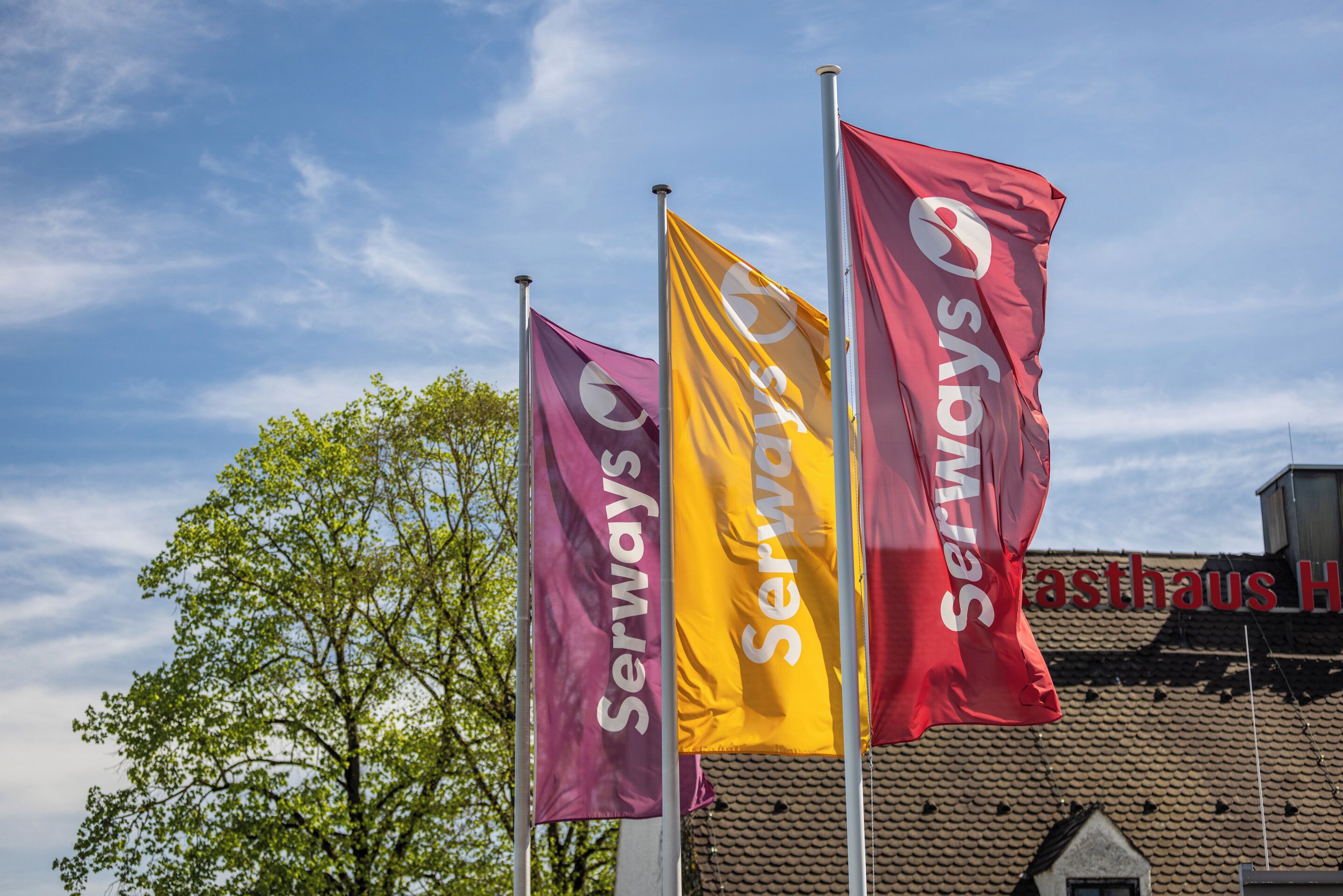Three flags with the word 'Serways' on them, a building with a tiled roof, and a tree in the background.