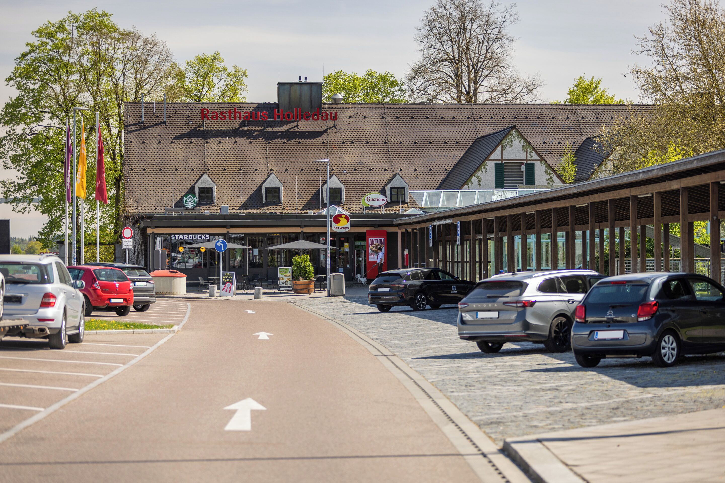 A parking lot with cars, a building with a sign reading 'Rasthaus Holledau,' and a storefront with 'Starbucks' and 'Gästehaus' signs.
