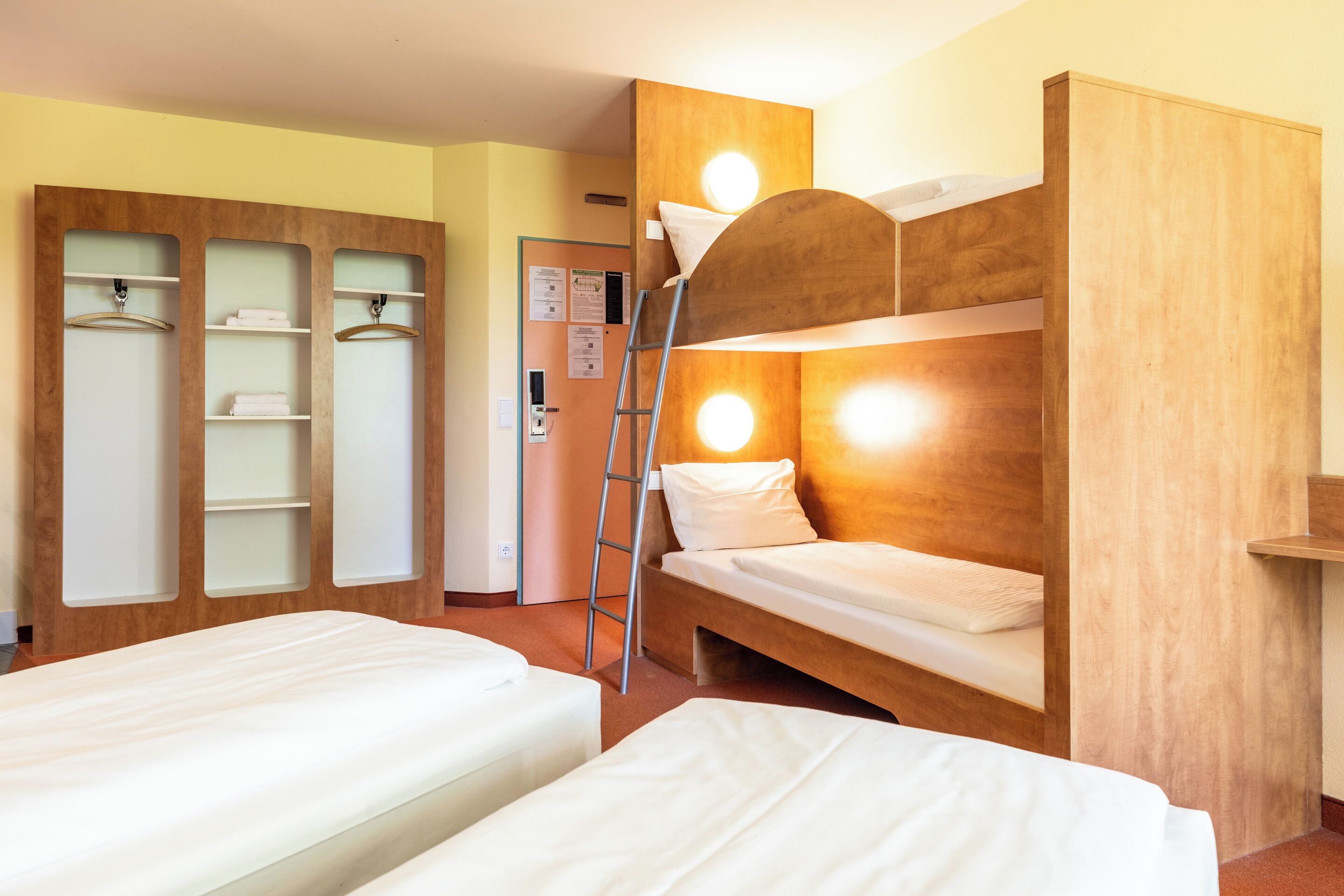 Een kamer met twee eenpersoonsbedden, een houten garderobe, een ladder en een deur met een prikbord.