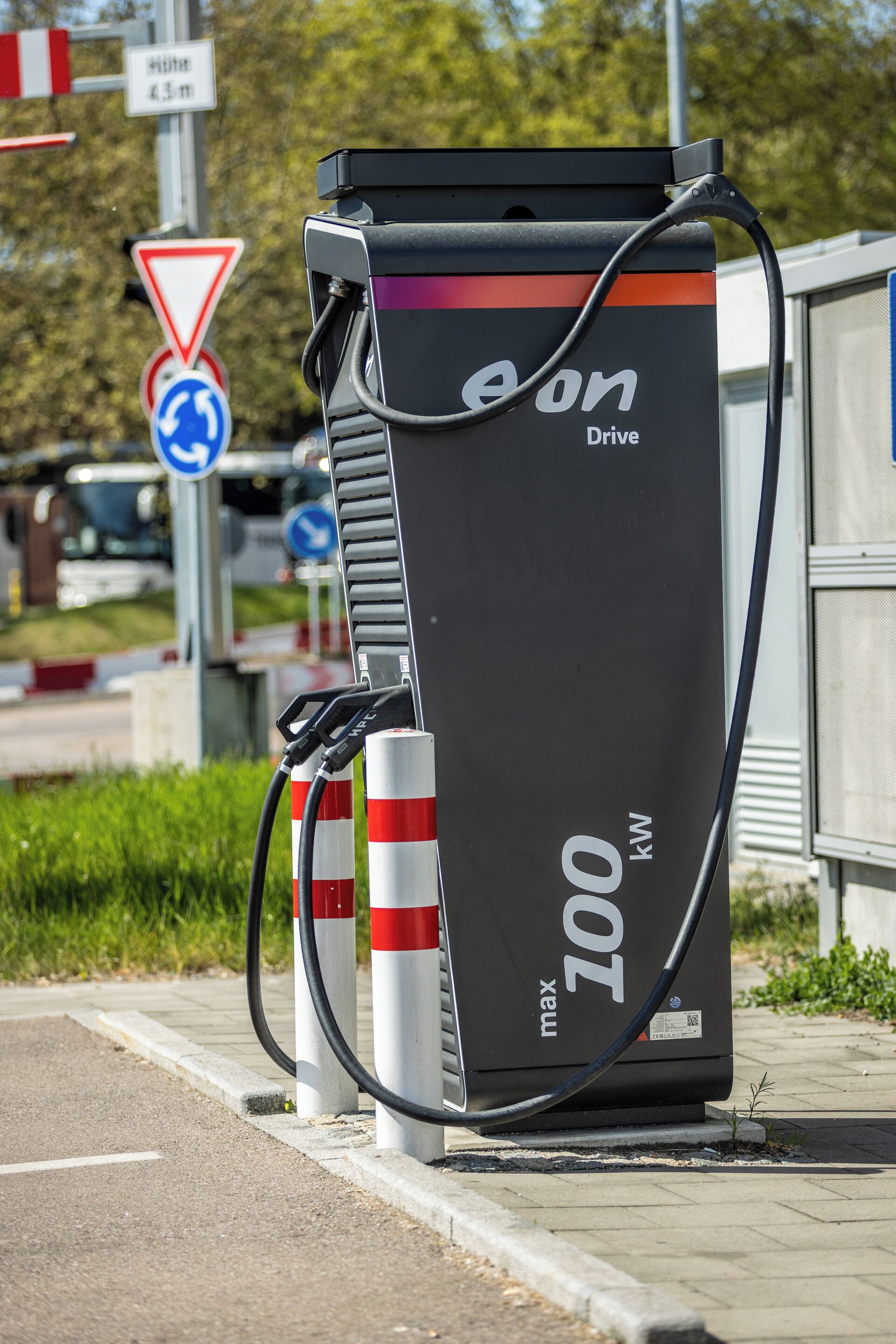 A 100 kW electric vehicle charging station with a black and red design, located on a paved roadside with traffic signs and greenery in the background.