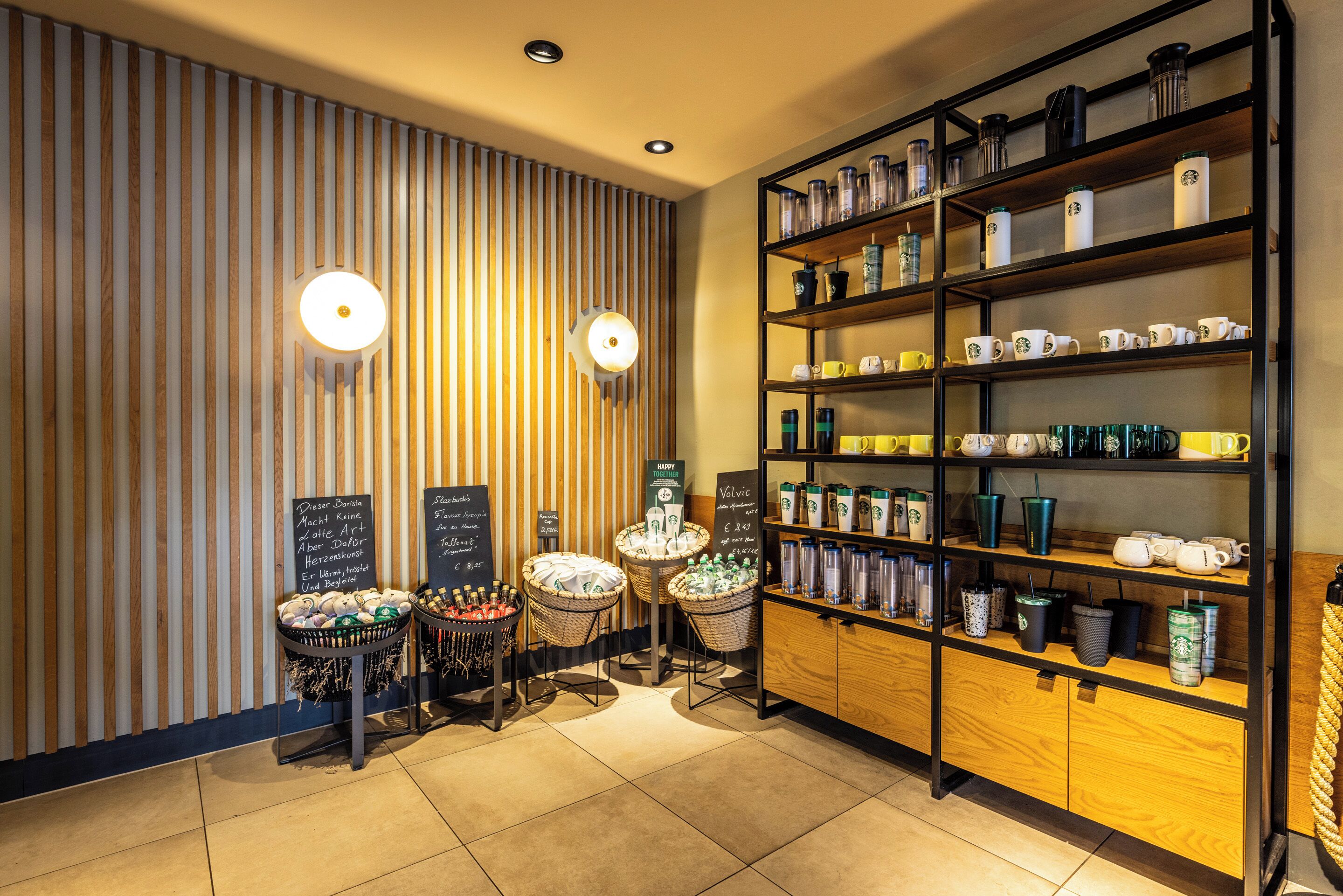 A café interior with wooden slats, shelves with mugs and cups, baskets with snacks, and chalkboard signs.