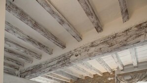Un dormitorio con vigas de madera a la vista, una pared de piedra, una cama con duvet gris, un televisor montado en la pared y una ventana con cortinas.