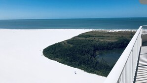 Ein weiter Blick von einer erhöhten Position auf einen Strand mit blauem Himmel, feinem Sand und bewaldetem Gebiet mit einem Gewässer.