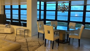 Ein modernes Wohnzimmer mit einem großen Fenster mit Meerblick, einem runden Glastisch mit Stühlen, einer Couch und einem TV-Schrank.
