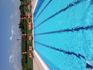 Una piscina con agua azul cristalina, rodeada de césped, sombrillas naranjas y un flotador salvavidas.