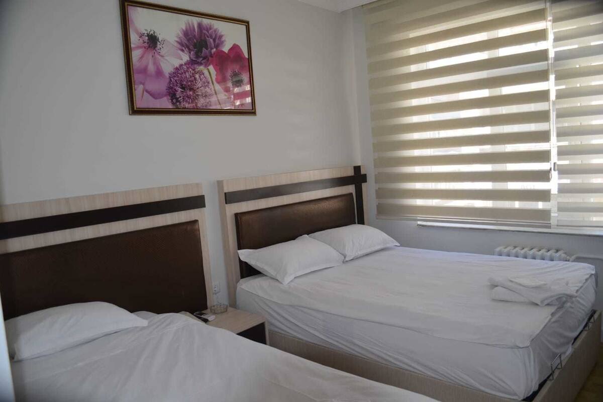 A hotel room with two beds, a nightstand, a framed floral artwork, and a window with blinds.
