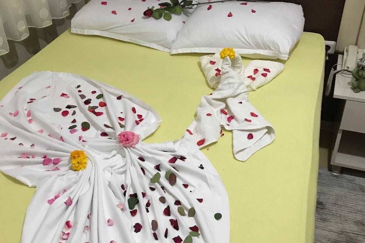 A hotel room with a yellow bedspread, white pillows, and a towel with rose petals and a yellow flower.