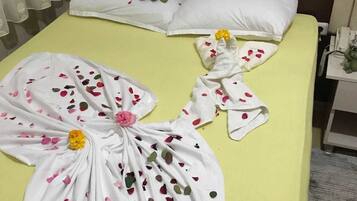 Une chambre d’hôtel avec une couvre-lit jaune, des oreillers blancs et une serviette décorée de pétales de rose et d’une fleur jaune.