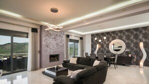 Ein modernes Wohnzimmer mit Sofa, Couchtisch und Kamin. Es gibt einen Essbereich mit Tisch und Stühlen, und ein großes Fenster mit Blick ins Freie.