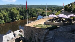 Vista al río con edificios de piedra, mesas al aire libre y banderas.