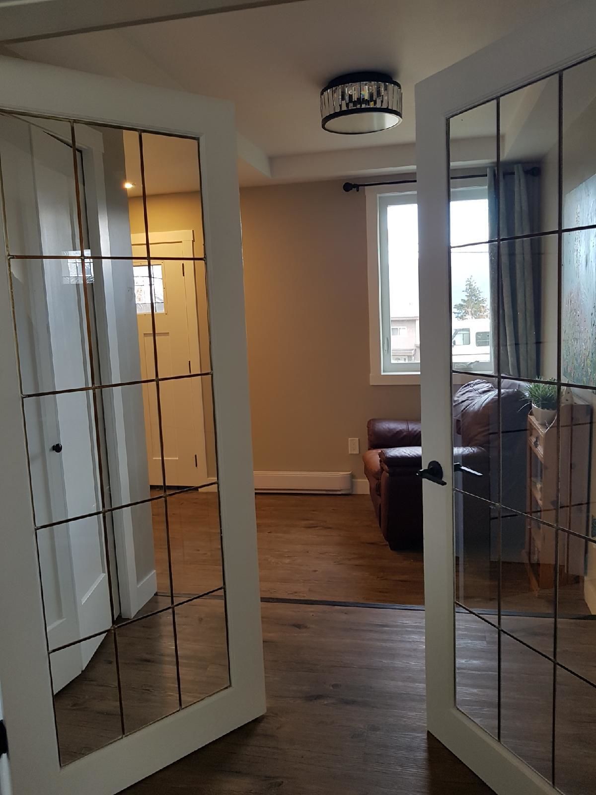Une pièce avec un plancher en bois, un canapé en cuir marron, une porte blanche à motifs géométriques et une fenêtre avec des rideaux.